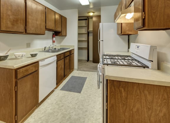 Narrow kitchen here at Taiga Apartments with wooden cabinets, beige countertops, a gas stove, white appliances, and a sink. A pantry, grey rug, and scattered bowls create a cozy feel.