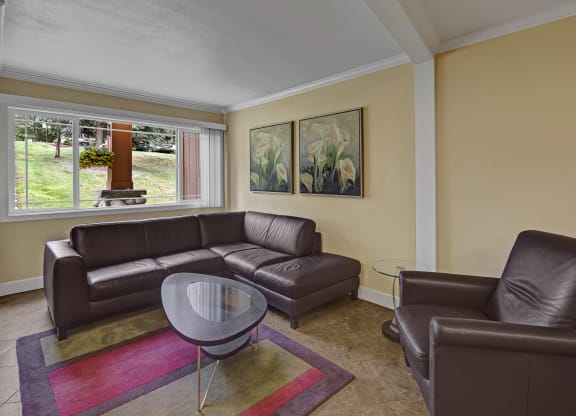 Cozy living room here at Taiga Apartments with brown leather sectional and armchair, a glass coffee table on a colorful rug, large window with garden view, and floral paintings on yellow walls.
