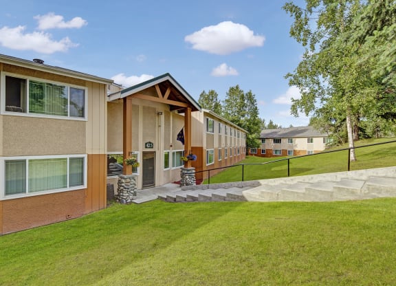 Two-story Taiga Apartment building with beige and orange siding, large windows, and a path leading up to the entrance. Lush green lawn and trees surround it.