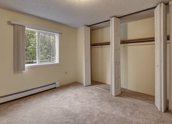 Empty carpeted bedroom here at Taiga Apartments with a large open closet featuring white folding doors. A window with blinds is on the left, revealing greenery outside. Neutral tones.