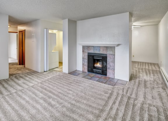 A spacious living room here at Terrace on the Lake Apartments with beige carpet features a lit fireplace with a tiled mantel. The room is bright, with white walls and a cozy feel.