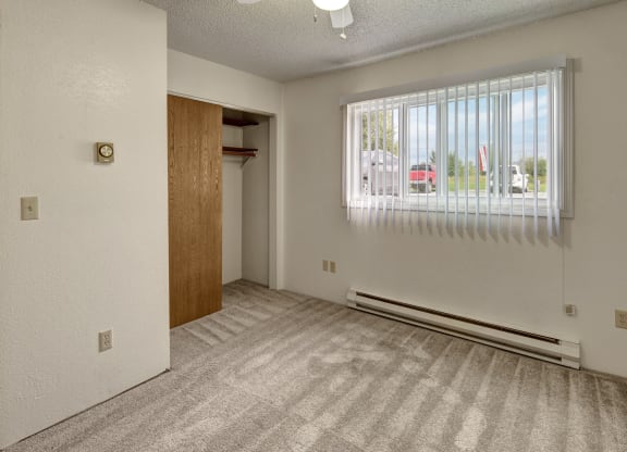 Empty bedroom Terrace on the Lake Apartments with beige carpet, white walls, and vertical blinds on a window. An open closet with a wooden door is on the left, creating a neutral, clean atmosphere.