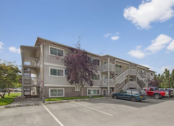 Three-story beige Terrace on the Lake Apartment building with external staircases, parked cars in front, and a cloudy blue sky. Trees frame the scene, offering a serene feel.
