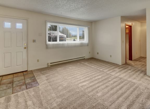 Empty living room here at Terrace on the Lake Apartments with beige carpet, white walls, and a light blue door with windows at the top. Large window with vertical blinds lets in natural light.