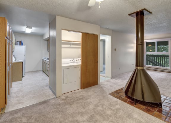 Open-plan living space here at Terrace on the Lake Apartments with beige carpet, a central vintage cone fireplace, and a windowed door leading to a balcony. Adjacent is a utility room with a washer and dryer.