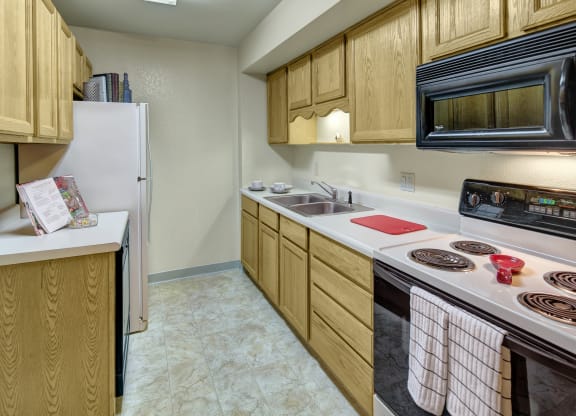 A narrow kitchen here at Terrace on the Lake Apartments with wooden cabinets, a double sink, stove, and microwave. A red cutting board and striped towels add a cozy touch. Bright lighting.