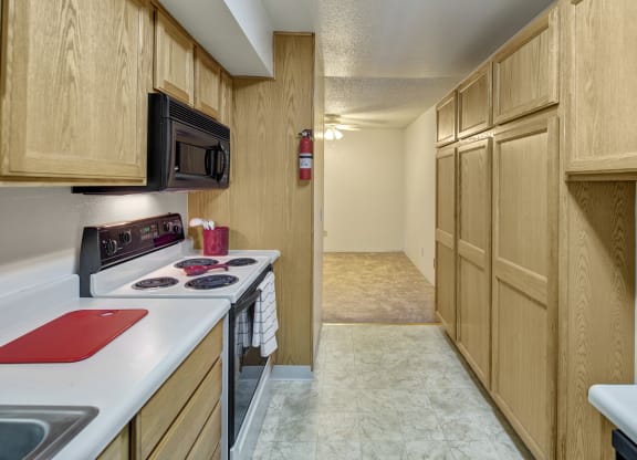 Narrow kitchen here at Terrace on the Lake Apartments with light wood cabinets, a black microwave, and a white stove. Red cutting board and utensils on counter. Bright, clean space.