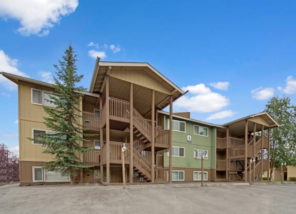 The Club at Eagle Pointe Apartments building with wooden exterior, green and beige tones. Three-story structure with staircases, surrounded by trees under a clear blue sky.