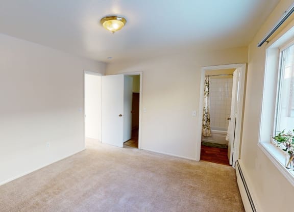 Empty bedroom here at the Village at Calais Apartments with beige carpet, cream walls, and a ceiling light. Open doors reveal a closet and a bathroom with a white tiled shower curtain. A large window adds brightness.