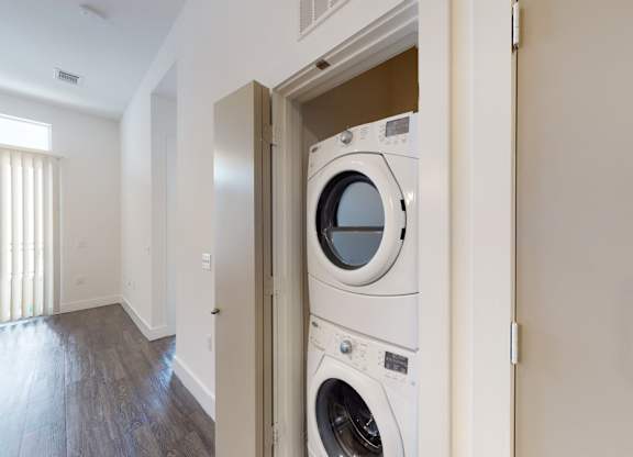 A convenient in-unit laundry area here at Arpeggio with a stacked washer and dryer set in a hallway closet, near bright living space with dark wood flooring and natural light.