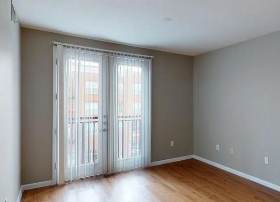 A bright empty living area here at BRIO at Firewheel with wood-style flooring, neutral walls, and a glass door with vertical blinds opening to a private balcony.