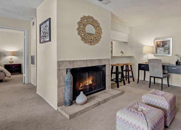 A cozy living room here at Cypress Pointe with soft beige carpet, neutral walls, and a lit fireplace creating a warm focal point. An open layout includes a built-in breakfast bar with seating and a small desk area, offering a comfortable and functional space for everyday living.