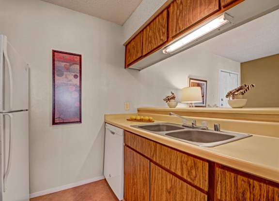A compact kitchen here at Cypress Pointe with warm wood cabinets, beige countertops, and a double stainless steel sink beneath bright overhead lighting. White appliances, including a refrigerator and dishwasher, add a clean, functional feel to this cozy, practical cooking space.