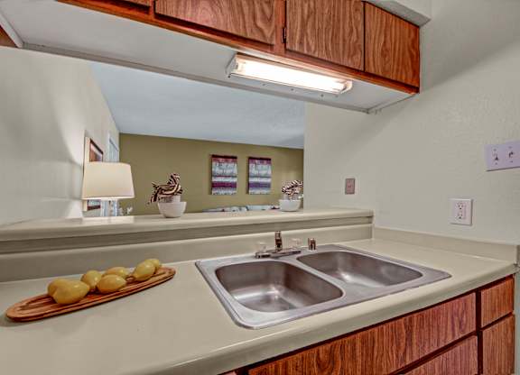 A functional kitchen here at Cypress Pointe with warm wood cabinets, beige countertops, and a double stainless steel sink beneath bright overhead lighting. An open pass-through overlooks the living area, creating a connected and practical layout for everyday use.