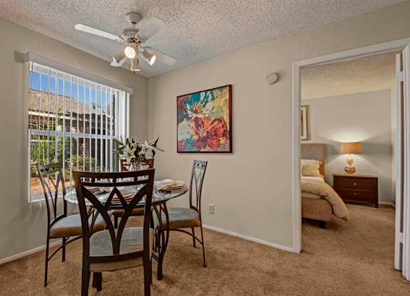 A bright dining area here at Cypress Pointe with beige carpet, neutral walls, and a large window with vertical blinds bringing in natural light. A ceiling fan adds comfort, while the open layout offers a clear view into the bedroom, creating a warm and inviting space for everyday dining.