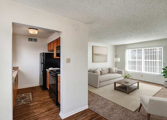 A bright, open living area here at Golden Crest with a cozy carpeted living room featuring a light neutral sofa, wooden coffee table, and large window with vertical blinds, connected to a compact kitchen with warm wood cabinets, dark appliances, and wood-style flooring that creates a clean, welcoming layout.