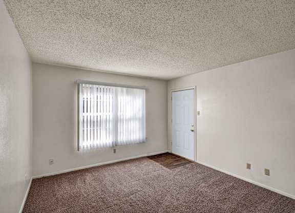 A bright, empty living room here at Golden Crest with plush carpeted flooring, soft neutral walls, and a large window with vertical blinds that lets in natural light, featuring a nearby entry door and a small section of wood-style flooring for a clean, open feel.