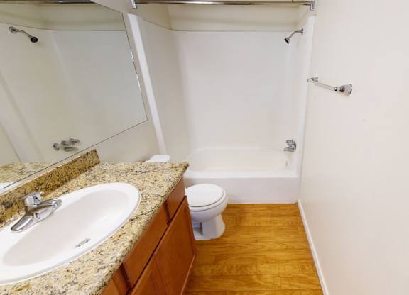 A clean, functional bathroom here at Golden Crest with a bathtub-shower combo, white toilet, and wood-style flooring, featuring a granite-style vanity countertop with a white sink and warm wood cabinetry for a bright, practical layout.