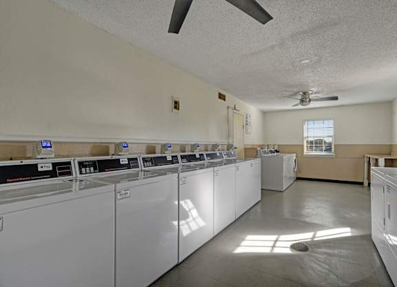 A bright, spacious laundry room here at Golden Crest with multiple white washers and dryers, smooth flooring, and ceiling fans, featuring large windows that bring in natural light for a clean and comfortable shared laundry space.