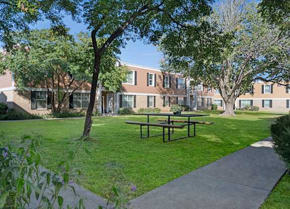 A quiet outdoor courtyard here at Golden Crest with a lush green lawn, shaded picnic table seating, and mature trees, surrounded by two-story apartment buildings and concrete walkways that create a relaxed, community-oriented setting.