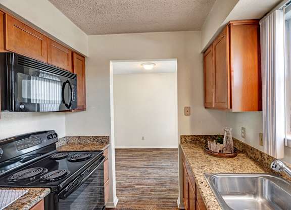 A galley-style kitchen here at Golden Crest with warm wood cabinets, granite-style countertops, a stainless steel sink, and black appliances, opening toward a nearby dining or living area with wood-style flooring and a bright, functional layout.