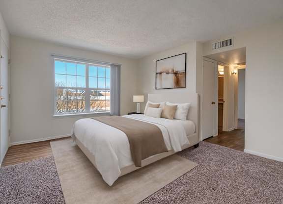 A bright, spacious bedroom here at Golden Crest with plush carpeted flooring, a neatly made bed in soft neutral tones, and a large window with vertical blinds that allows natural light to fill the room, creating a calm and comfortable retreat with easy access to the adjoining hallway and bathroom.