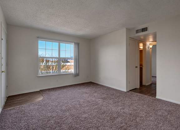 A spacious, empty living room here at Golden Crest with plush carpeted flooring, soft neutral walls, and a large window with vertical blinds that lets in natural light, offering an open layout with easy access to the hallway and adjacent rooms.