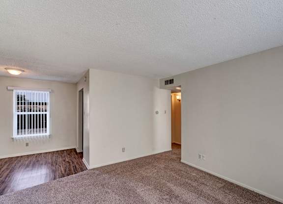 A spacious, empty living area here at Golden Crest with plush carpeted flooring, soft neutral walls, and a large window with vertical blinds, featuring an open layout that connects to a wood-style flooring area and provides easy access to the hallway.