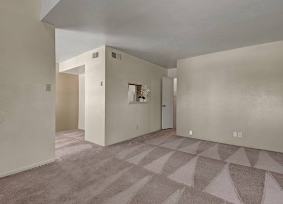 A spacious, empty living room here at High Plains with soft beige carpet, neutral walls, and an open layout, featuring a pass-through window to the kitchen and multiple wall openings that create a bright, flexible space for everyday living.