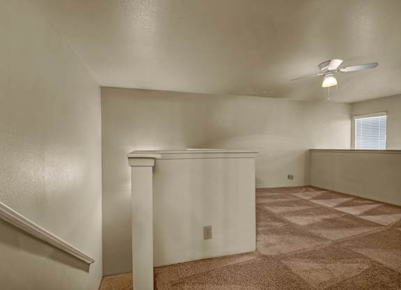 A spacious upstairs loft here at High Plains with soft beige carpet, neutral walls, and a ceiling fan with built-in lighting, featuring an open railing overlook and natural light from a window that creates a flexible, comfortable bonus living space.