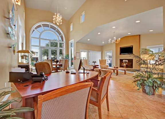 A welcoming community lounge here at High Plains with a shared work table, comfortable seating, tall arched windows, and a cozy fireplace with a mounted TV, enhanced by warm lighting and an open layout that creates a relaxed, social atmosphere.