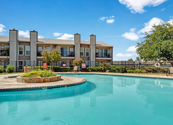 Here at High Plains, a bright outdoor swimming pool with clear blue water sits in a landscaped courtyard, framed by mature trees, fencing, and surrounding apartment buildings with balconies under a sunny blue sky.