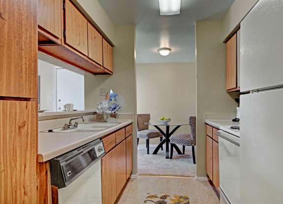 A narrow kitchen here at High Plains with warm wood cabinets, light countertops, and white appliances, featuring a dishwasher and a compact layout that opens to a small dining area, creating a simple, functional, and comfortable cooking space.