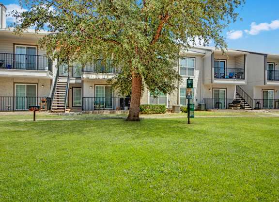 A well-maintained outdoor lawn here at High Plains with lush green grass, a large shade tree, and apartment buildings featuring private patios and balconies. A small community grill and open green space create a relaxed, inviting area for outdoor enjoyment.