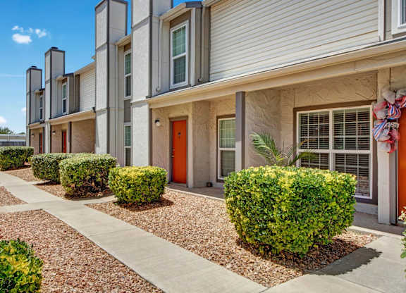 A clean exterior walkway here at High Plains with neatly landscaped shrubs, desert-style gravel beds, and townhouse-style apartment entrances with bright accent doors. The building features large windows and a tidy, welcoming layout under clear blue skies.