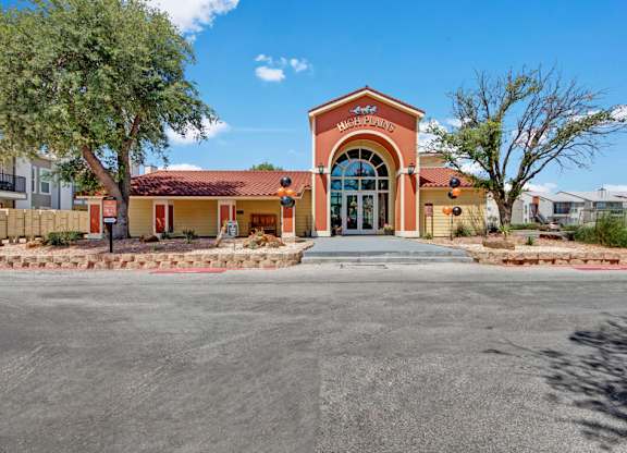 A welcoming leasing office exterior here at High Plains with a charming entryway, arched glass doors, and warm-toned stucco accents. Desert landscaping, mature trees, and a tiled roof create a polished and inviting first impression for residents and guests.