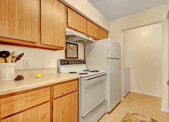 A compact kitchen here at High Plains with warm wood cabinets, light countertops, and white appliances, including a stove and refrigerator, offering a simple, functional layout with a clean and cozy feel.