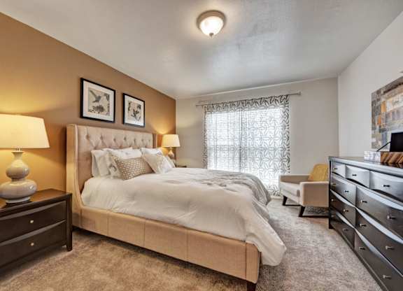 A welcoming bedroom here at High Plains with plush beige carpet, a neutral upholstered bed, warm accent walls, and soft lighting, complemented by a large window with patterned curtains and tasteful furnishings that create a calm, comfortable retreat.