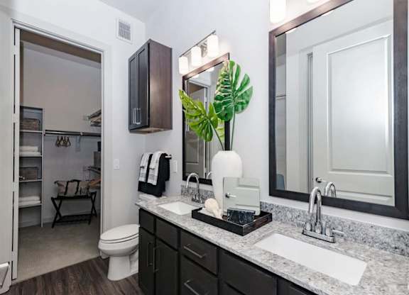 A modern bathroom here at Magnolia on the Green with a double-sink vanity, granite countertop, dark cabinets, framed mirrors with bright lighting, and an open walk-in closet with built-in shelving nearby.