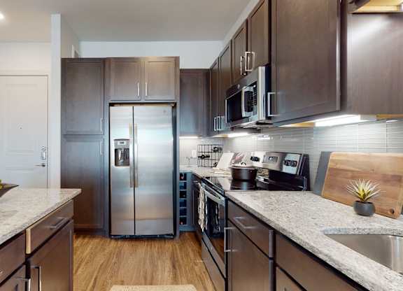 A sleek kitchen here at Magnolia on the Green with dark wood cabinets, granite countertops, stainless steel appliances, under-cabinet lighting, and a large island, creating a bright, modern cooking space.