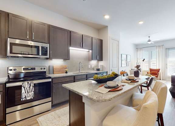 A bright open kitchen and living area here at Magnolia on the Green with dark cabinets, granite countertops, stainless steel appliances, an island with seating, wood-style floors, and large glass doors bringing in natural light.