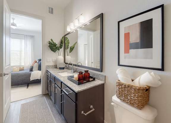 A bright bathroom here at Magnolia on the Green with a dark wood vanity, granite countertop, wide mirror with modern lighting, and stylish decor, connected directly to a nearby bedroom for convenient access.