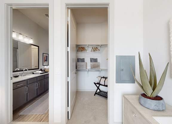 A convenient bedroom hallway here at Magnolia on the Green with plush carpet, an open walk-in closet with built-in shelving, and a nearby bathroom vanity with granite countertop and bright lighting.