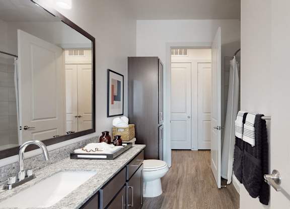 A clean bathroom here at Magnolia on the Green with a dark wood vanity, granite countertop, large mirror, tile shower, and wood-style flooring, creating a bright, modern, and functional space.