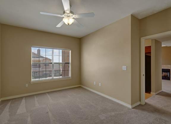 A spacious bedroom here at Mission Green featuring soft neutral walls, plush carpet flooring with clean vacuum lines, a ceiling fan with overhead lighting, and a large window with blinds that brings in natural light, creating a comfortable and versatile space with an open layout that connects easily to the adjacent living area.
