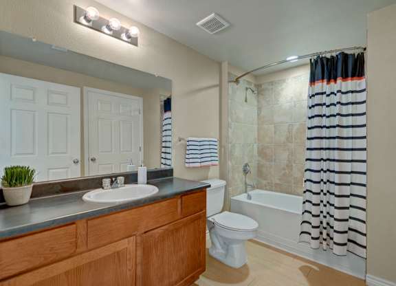 A bright bathroom here at Mission Green showcasing a wide vanity with a wood-tone cabinet, a large wall mirror with overhead lighting, a clean tub and shower combo with tile surround, light neutral walls, and coordinated fixtures that create a fresh, functional, and well-lit space designed for everyday comfort and convenience.