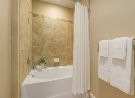 A clean and inviting bathroom here at Mission Green featuring a spacious tub with tiled surround, a crisp white shower curtain, neatly folded white towels on a wall-mounted rack, warm neutral wall tones, and simple decorative accents that create a bright, comfortable, and well-maintained bathing space.