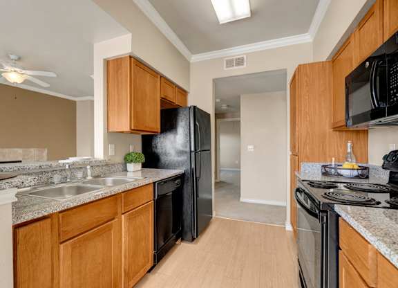 A warm and functional kitchen here at Mission Green featuring wood-style cabinetry, speckled countertops, a double-basin sink, and full-size appliances arranged along a galley layout, with durable flooring, overhead lighting, and an open view toward the living area that supports easy cooking and everyday use.