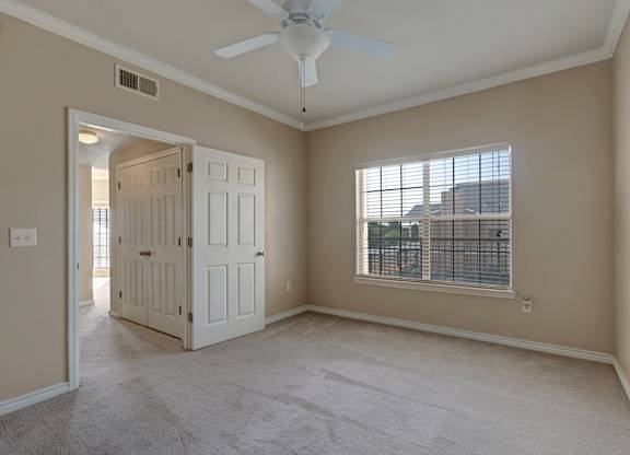 A comfortable bedroom here at Mission Green featuring soft neutral walls, plush carpet flooring, a ceiling fan with overhead lighting, and a large window with blinds that brings in natural light, with double-door closet access and an open doorway that connects conveniently to the rest of the apartment.