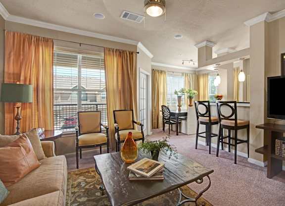 A warm, inviting living room here at Palms at Briarwood featuring plush carpeting, a comfortable sofa with accent pillows, a rustic coffee table, and soft window treatments that frame a private balcony, with an open layout flowing into a dining area and breakfast bar with barstools under modern pendant lighting.